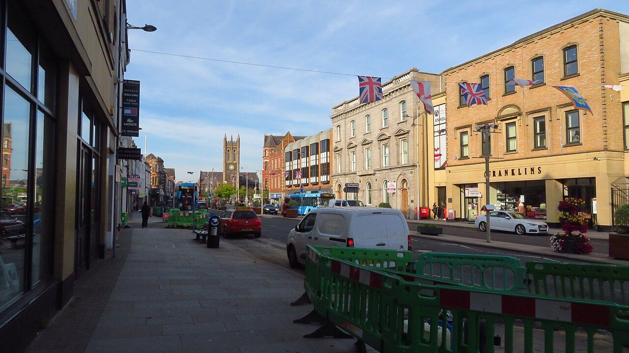 Portadown High Street with shopfronts, flags, a parked van and green roadworks barriers