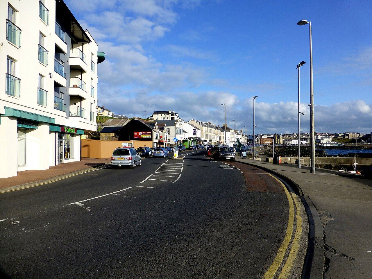 The Promenade in Portstewart with parked cars, seafront flats and shops curving round towards the harbour