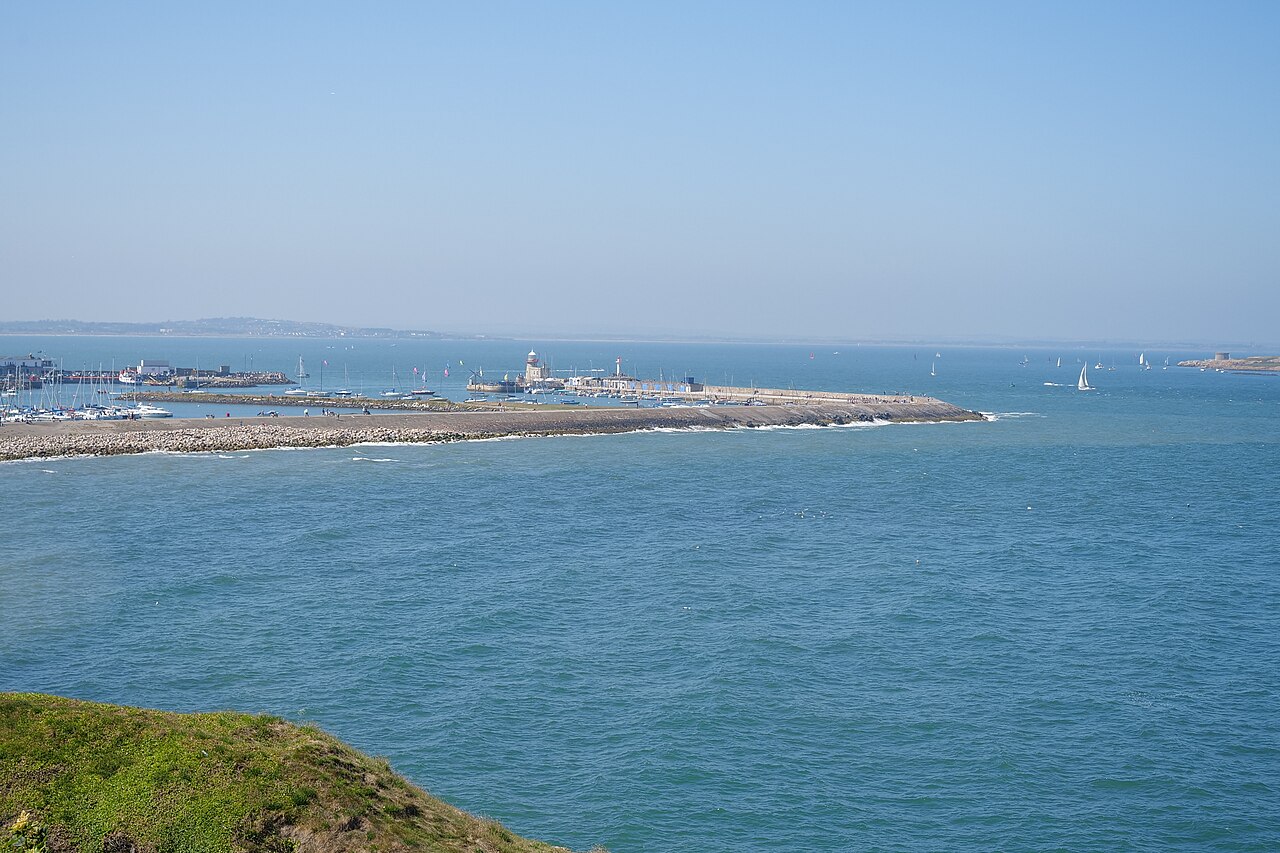 Howth Harbour's pier and lighthouse across calm blue water, with boats in the marina.