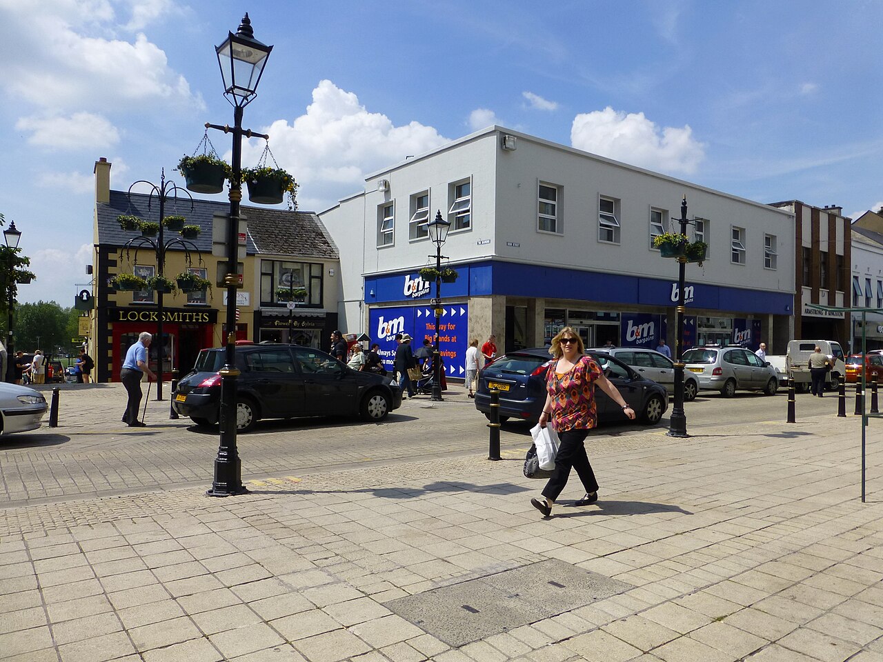 Paved street in Enniskillen town centre with an iron lamp post, parked cars, shopfronts and people walking past