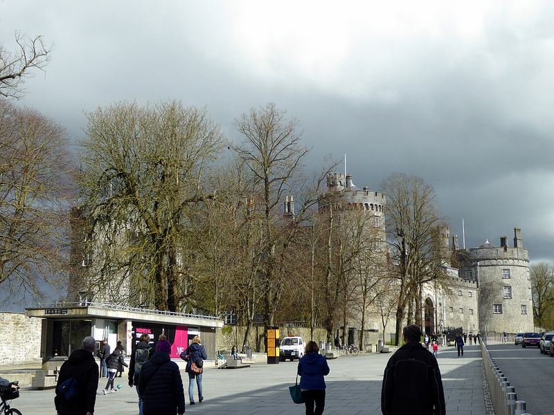 Kilkenny Castle's round towers beyond bare trees on The Parade, people walking under a grey sky