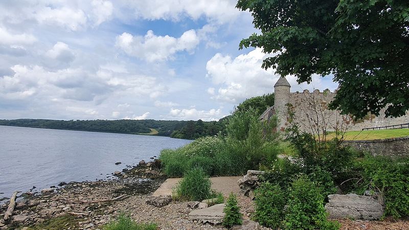 Stony shore of Lough Gill with the walls and corner tower of Parke's Castle behind trees