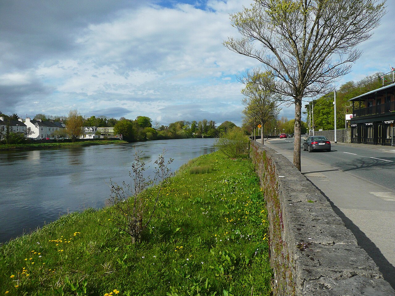 The River Moy in Ballina beside a stone riverside wall and road, houses on the far bank