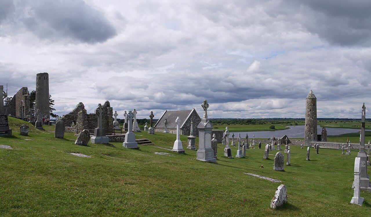 Round tower, ruined churches and high crosses at Clonmacnoise under a grey sky, the Shannon behind