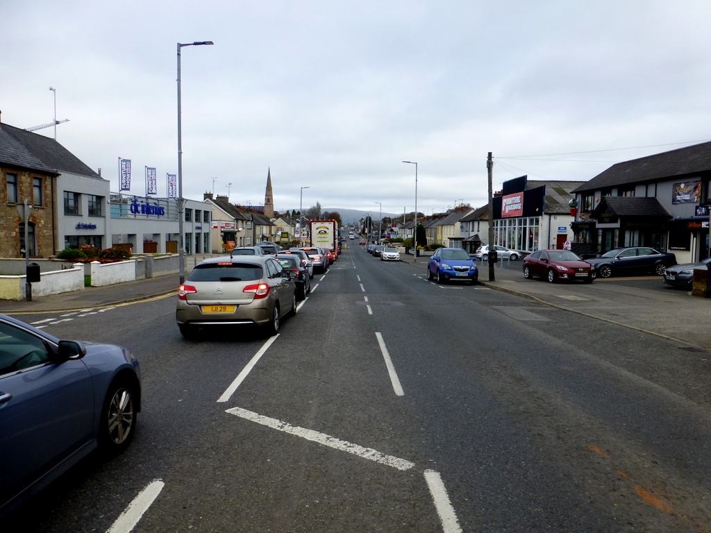 Looking up Cookstown's wide main street past parked cars and shops towards a church spire on an overcast day