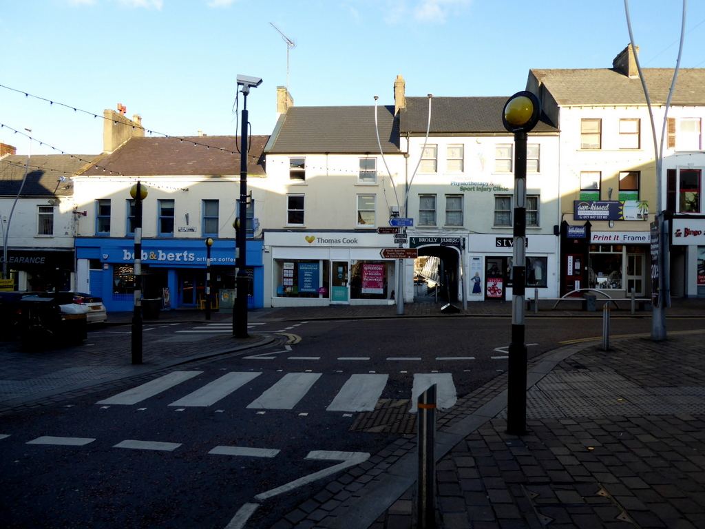 Pedestrian crossing at the foot of High Street in Omagh, with a row of shopfronts and a travel agent in low sun