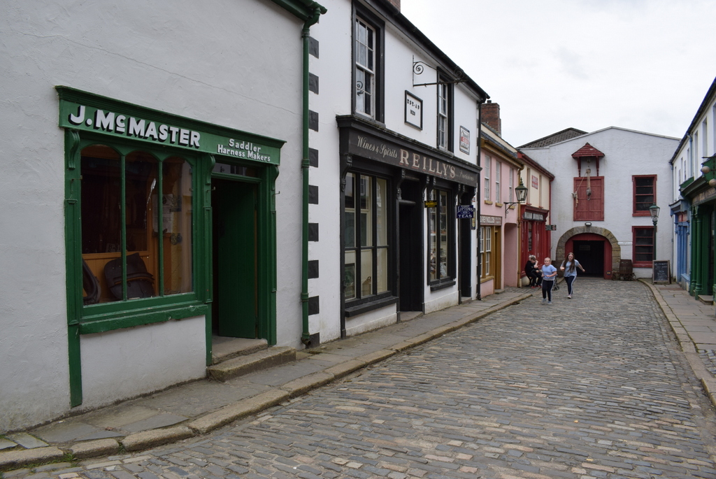 Cobbled street of rebuilt shopfronts at the Ulster American Folk Park, with a saddler's shop and a few visitors